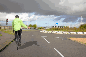 <p>You're approaching this roundabout. What should you do when a cyclist is keeping to the left while signalling to turn right?</p>