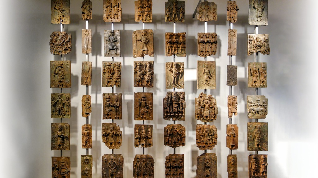 <p>A brass plaque from the Benin Kingdom shows a royal figure larger than attendants, richly adorned, and surrounded by symbolic motifs. Which is a key function of royal arts like this?</p>